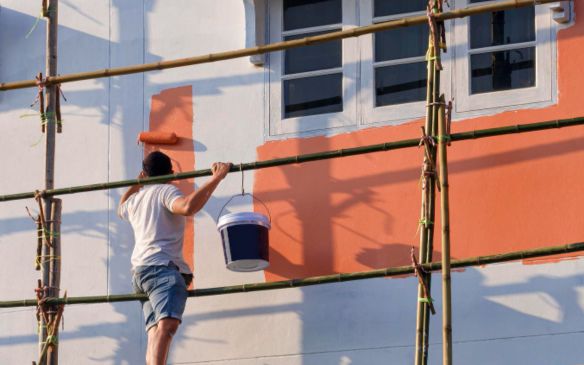 a man painting a house with orange paint, holding a paintbrush and wearing work clothes.